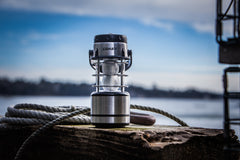 A COAST stainless steel lantern sitting on a wooden dock next to a rope on a sunny day with the ocean in the background. 