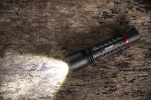 An illuminated LED flashlight sitting next on top of a textured aluminum surface next to piping and a pressure gauge. 