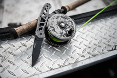 An open folding knife with a black stainless steel blade leaning against a fishing lure with a neon green fishing line.