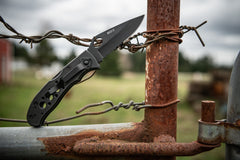 Stainless Steel Folding Knife Resting against Wire Fence, lifestyle photo.