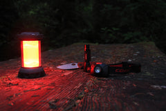 An illuminated red light LED lantern on a wooden park bench next to a folding knife, flashlight, and headlamp. 