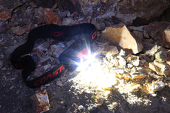 An LED headlamp sitting on gravel surrounded by rocks turned on to white light mode.