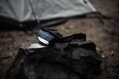 An illuminated COAST FL13 LED Headlamp sitting on a rock next to a gray tent.  