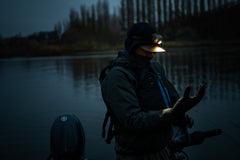 A man standing on a boat in a river using an LED headlamp to put on his waterproof glove. 