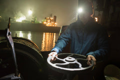 A man on a tugboat using an LED headlamp to turn a circular lever at night on a river.