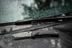 A black stainless steel folding knife with a seat belt cutter and glass breaker sitting on top of a wet car hood. 